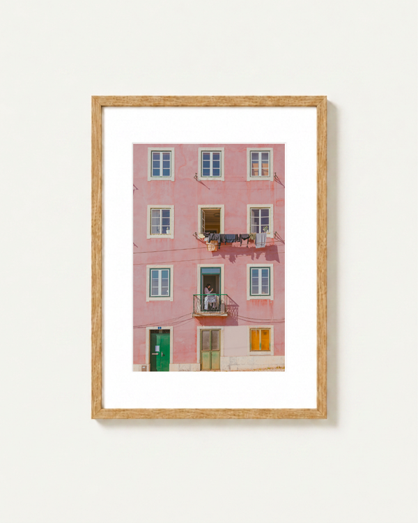 Framed wall art featuring a photograph of a pink apartment building facade in Lisbon with white-framed windows, a green door, and small balcony details, displayed in a light wood frame against a white wall.