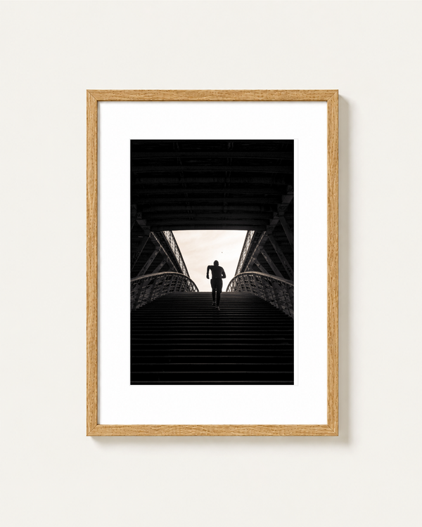A framed black and white art print featuring a runner's silhouette on a bridge staircase, set in a light wood frame with a white mat against a light beige wall, photographed by Stef Leny in Paris.
