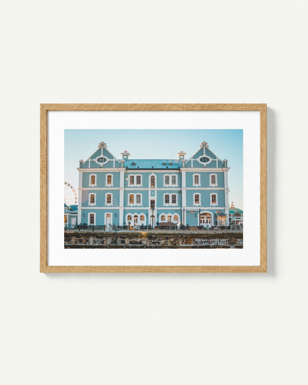A framed art print featuring a light blue seaside building in Cape Town with white trim, displayed on a clean off-white wall. The print is matted in white and enclosed in a natural light wood frame.