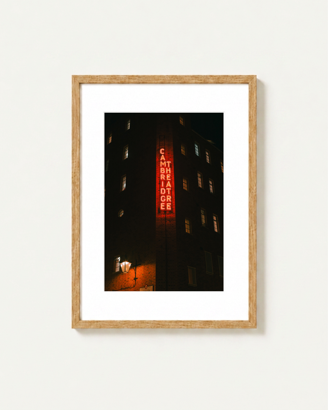 Framed vertical art print of the Cambridge Theatre in London at night, featuring a glowing red neon sign against a dark building facade, presented in a light wood frame with a white mat on a light gray background.