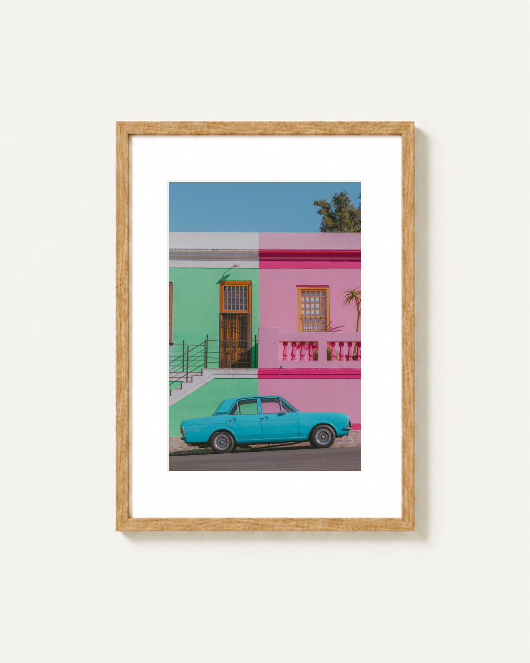 A framed art print featuring a colorful street scene in Cape Town with a turquoise car parked in front of a split green and pink building under a blue sky, displayed on a light wall.