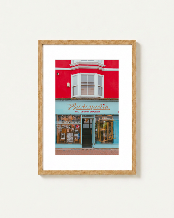 A framed art print depicting a vibrant red building facade in Brighton with a turquoise storefront sign reading Photomatic Photobooth Emporium, shown against a clean white wall.
