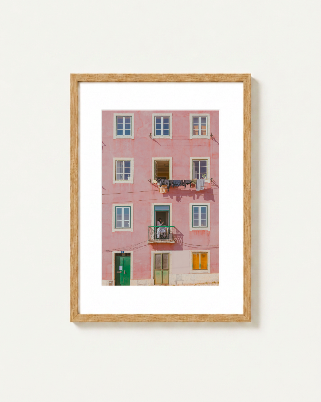 Framed wall art featuring a photograph of a pink apartment building facade in Lisbon with white-framed windows, a green door, and small balcony details, displayed in a light wood frame against a white wall.