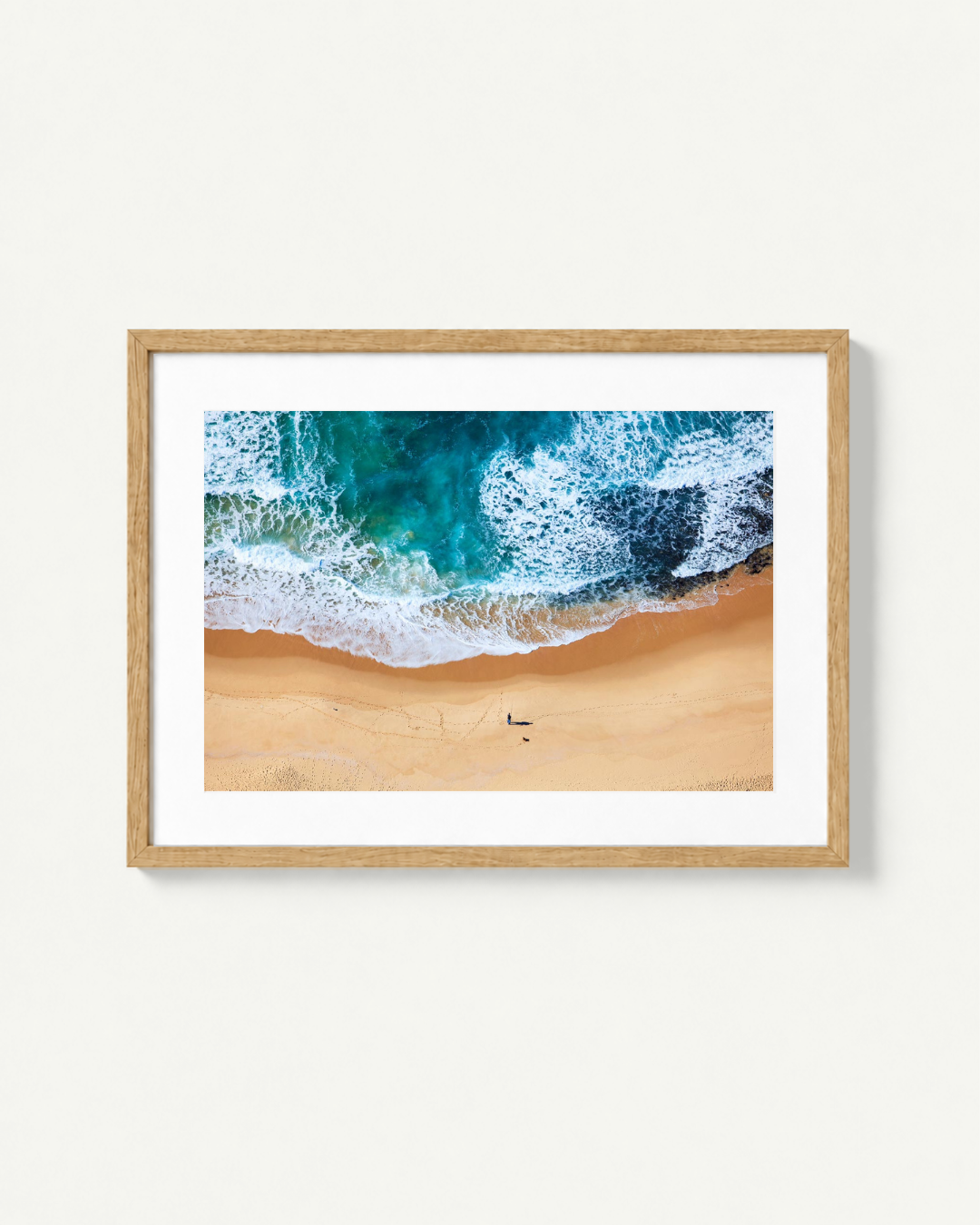 A framed aerial photograph of turquoise ocean waves crashing onto a sandy beach, presented in a light wood frame with a white mat against a plain off-white wall.