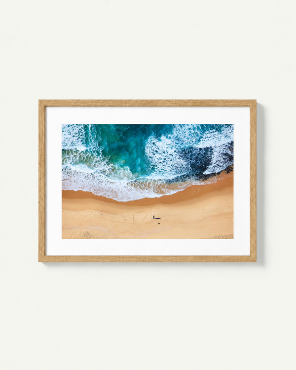A framed aerial photograph of turquoise ocean waves crashing onto a sandy beach, presented in a light wood frame with a white mat against a plain off-white wall.