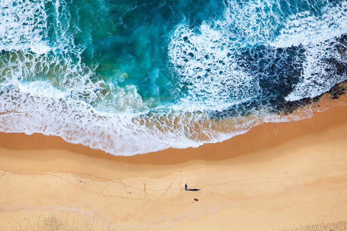 Aerial photograph of turquoise ocean waves meeting a sandy beach with a lone fisherman walking along the shoreline, photographed by Richard du Toit