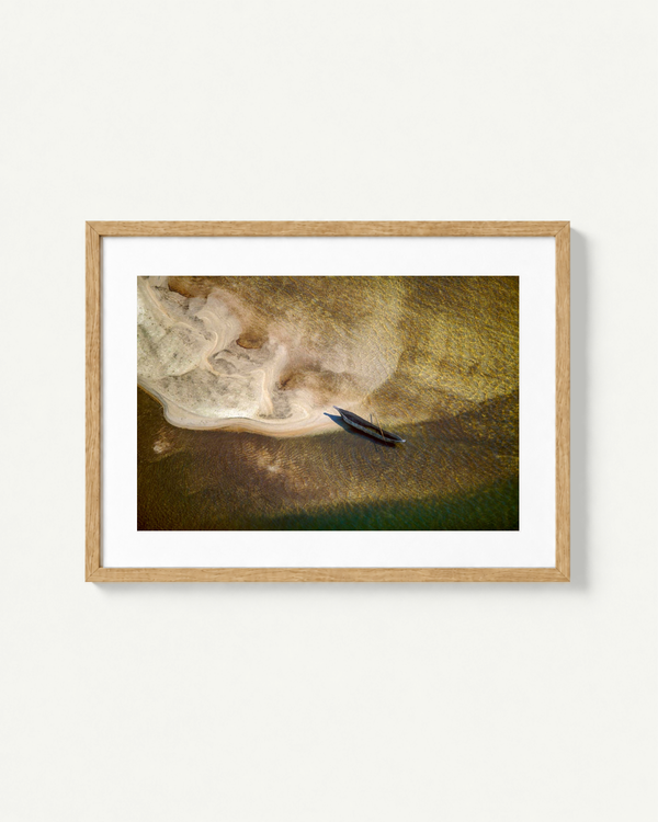 A framed art print featuring an aerial photograph of a mokoro boat in shallow water, presented in a light oak wood frame with a white mat against a clean white background.