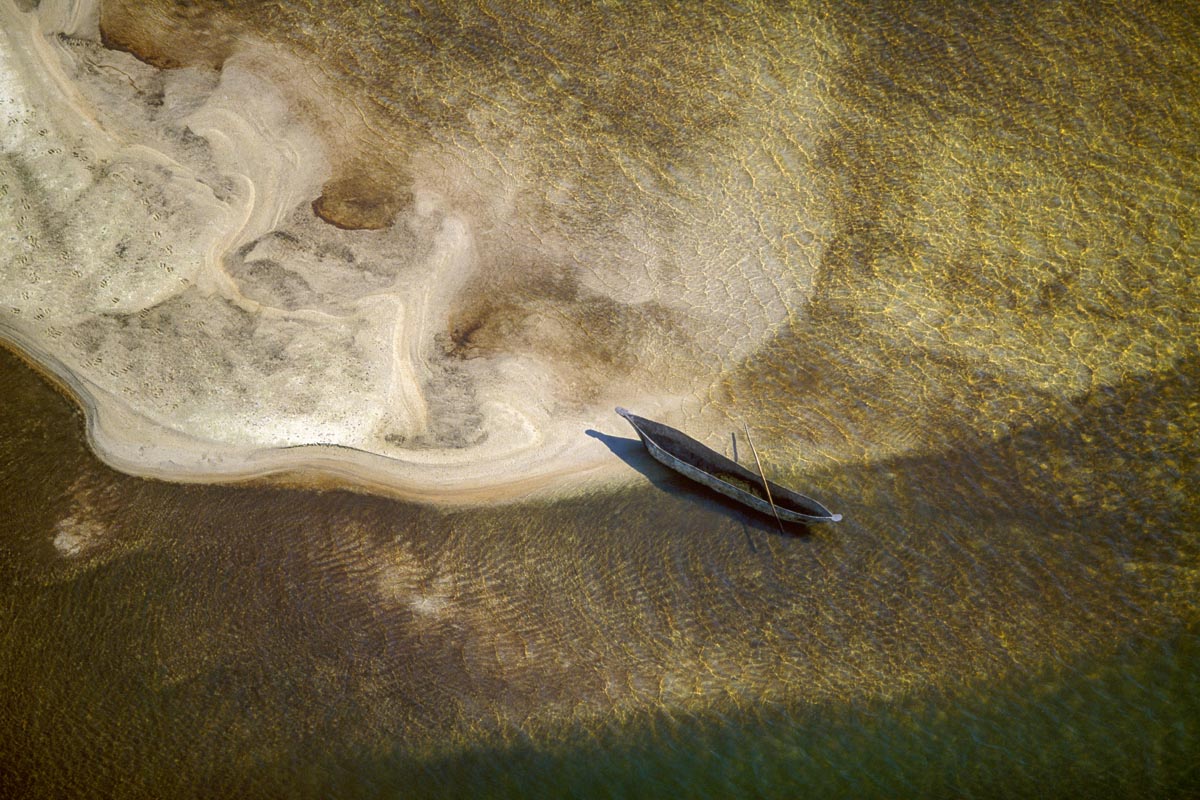 Aerial photograph of a small African wooden mokoro boat resting along the edge of a shallow sandy shoreline surrounded by rippling golden water, photographed by Richard du Toit