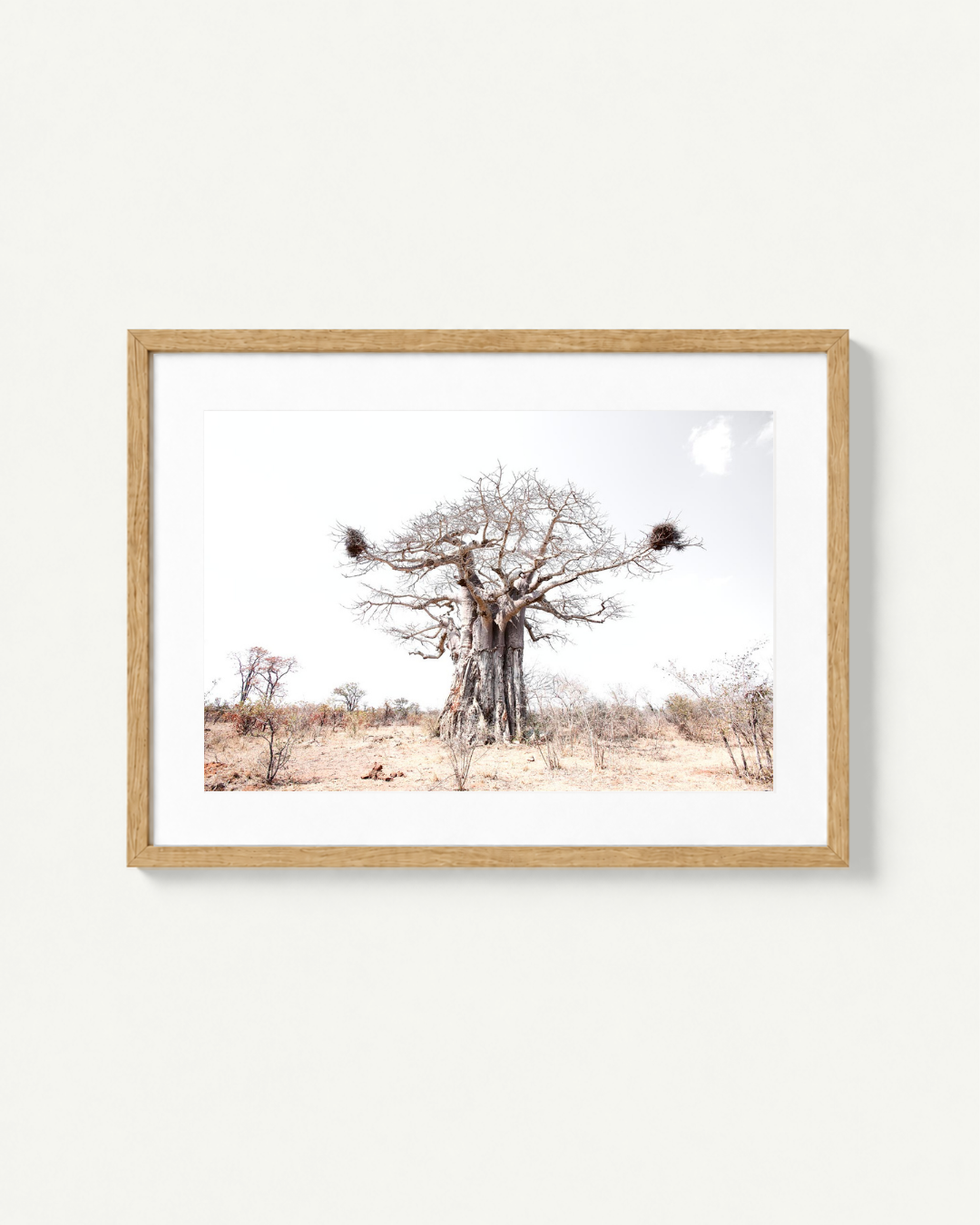 A framed art print depicting a desert landscape with a large leafless tree, sparse shrubs, and bird nests on its branches, set in a light wood frame against a white background.