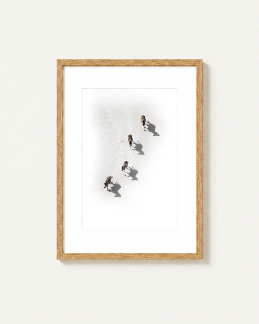 A framed art print featuring an aerial photograph of four zebra walking in a bright landscape, presented in a light natural wood frame with a white mat against a clean white wall.