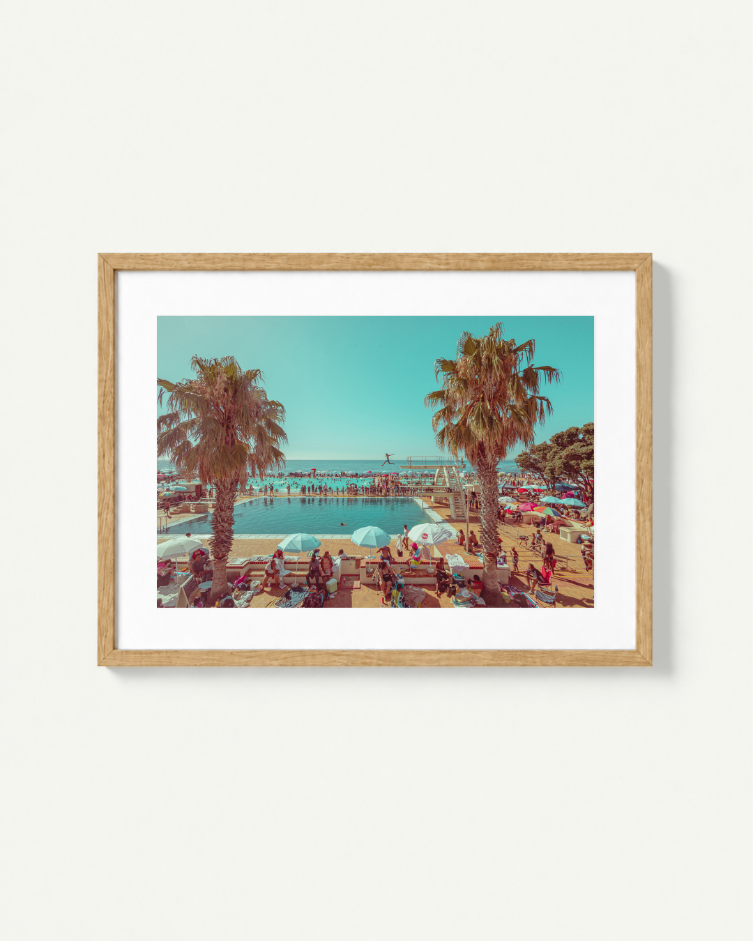 A framed art print featuring a retro-style photograph of a beach scene in Cape Town with palm trees, poolside loungers, and a pier under a clear blue sky, displayed on a light beige wall.