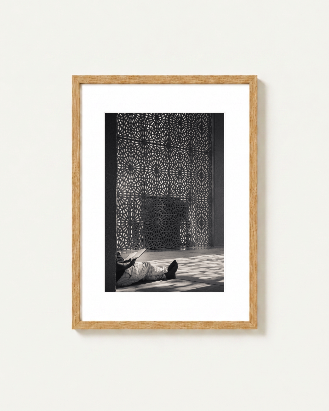 A framed black and white art print featuring a person lying on a floor in a place of worship in front of an intricate geometric screen, set in a light wood frame with a white mat against a light gray wall.