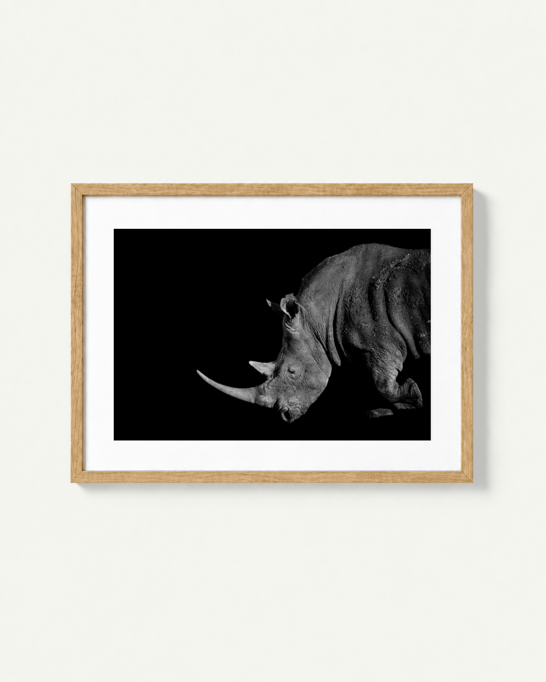 Framed black and white photographic print of a rhinoceros, shown in a light wood frame with a white mat against a clean off-white wall.