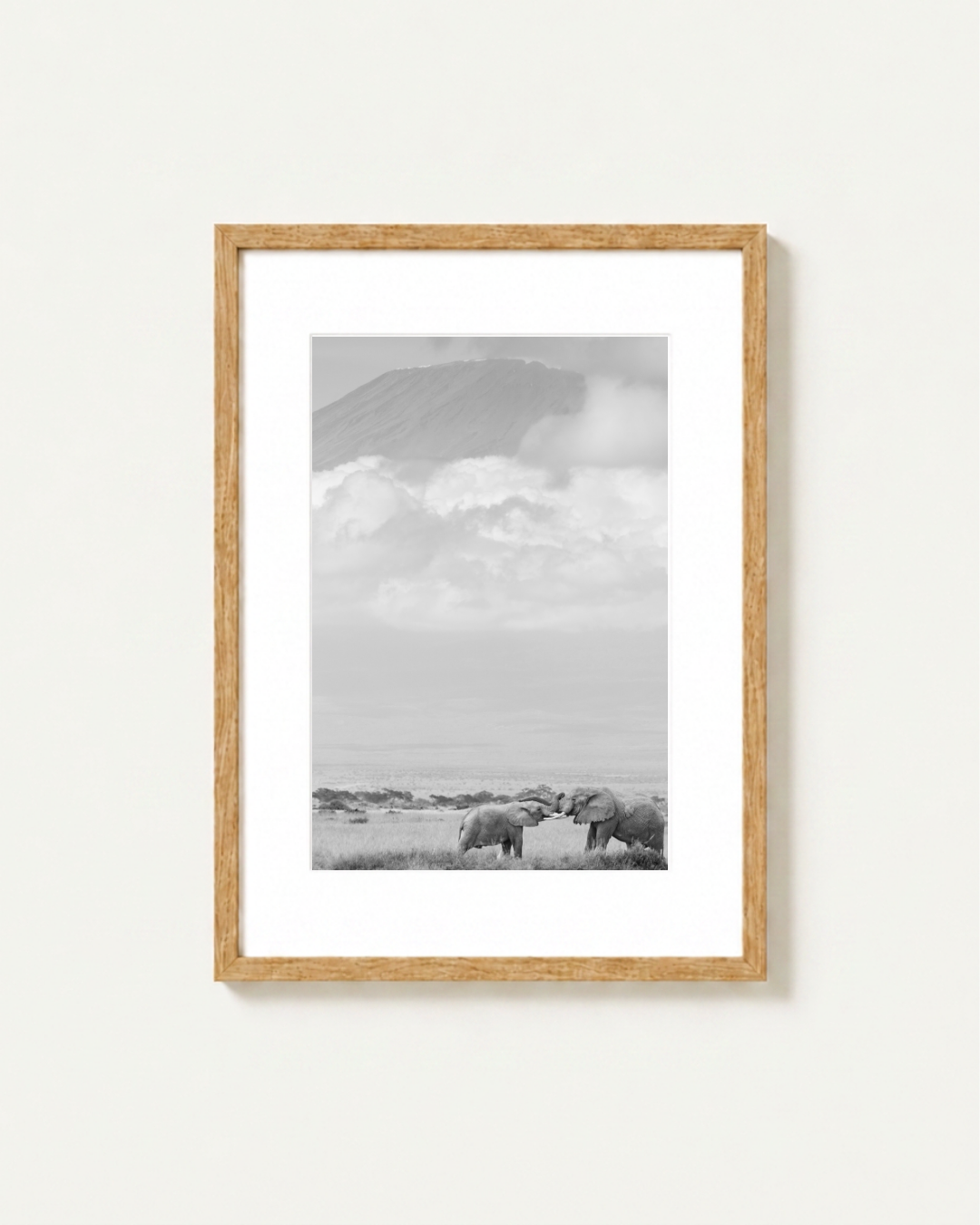 A framed black and white landscape photograph featuring elephants in Amboseli under a cloudy sky with the Kilimanjaro in the background, presented in a light wood frame with a white mat on a light beige wall.
