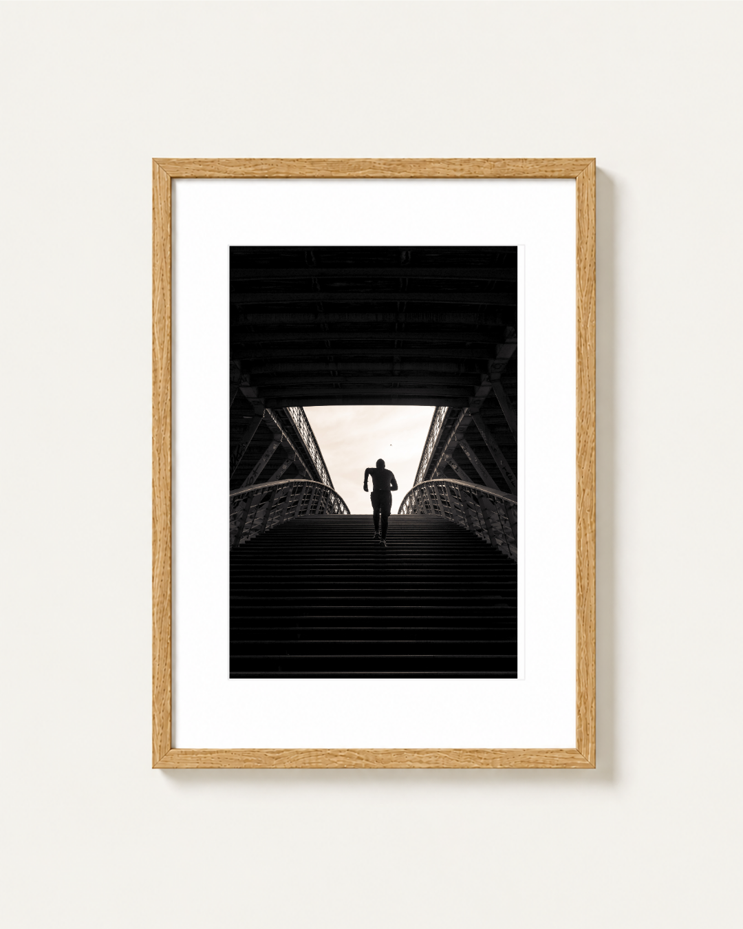 A framed black and white art print featuring a runner's silhouette on a bridge staircase, set in a light wood frame with a white mat against a light beige wall, photographed by Stef Leny in Paris.